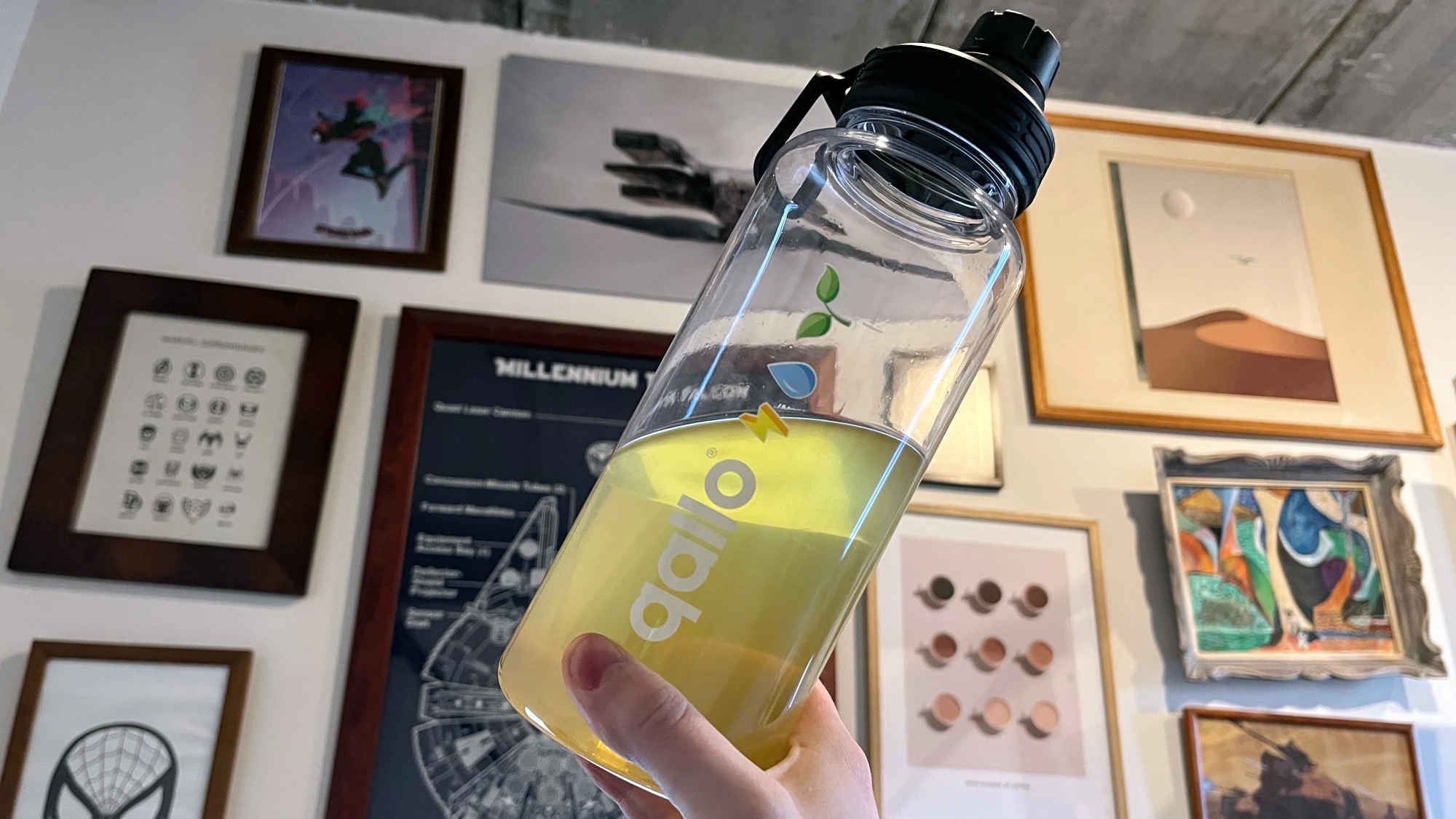 Een foto van een Qallo-drinkbus, met een lichtgele vloeistof erin. Op de achtergrond zien we verschillende kaders met afbeeldingen van Spider-Man, Marvel, Star Wars en andere kunstzinnige objecten.